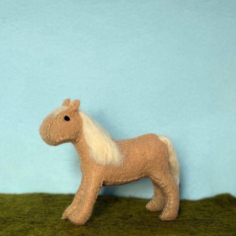 May include: A handmade felt horse toy in shades of tan and cream. The horse has a cream-colored mane and tail, with a stitched design. The toy is standing on a green surface, with a light blue background.
