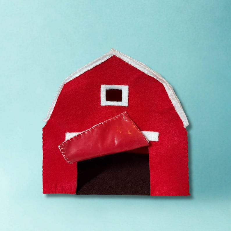 May include: A red felt barn with a white roof and trim. The barn door is open, revealing a dark brown interior. A small white square window is visible near the roofline. The barn is set against a light blue background.