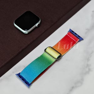 Puede incluir: Una correa de reloj multicolor con conectores azules y una hebilla plateada. La correa hace la transición del azul al verde, amarillo, naranja y rojo. Un Apple Watch plateado también está en la imagen.