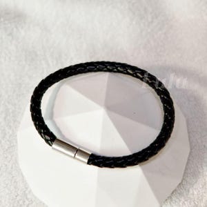 May include: A black braided leather bracelet with a silver magnetic clasp. The bracelet is circular and sits on a white geometric surface. The leather is tightly woven, and the clasp is a bright silver color.
