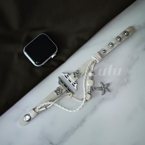 May include: Cream-colored Apple Watch band with silver-tone hardware and a starfish charm. The band features decorative metal studs and a braided rope design. The watch face is black with a silver-tone frame. The word "lulu" is printed on the band.