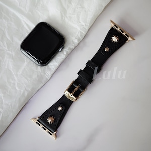 May include: Black leather watch band with gold-colored hardware, including a buckle and starburst accents. The band is shown next to a black smartwatch with a dark screen. The word "Lulu" is printed on the band.