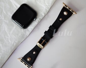 May include: Black leather watch band with gold-colored hardware, including a buckle and starburst accents. The band is shown next to a black smartwatch with a dark screen. The word "Lulu" is printed on the band.