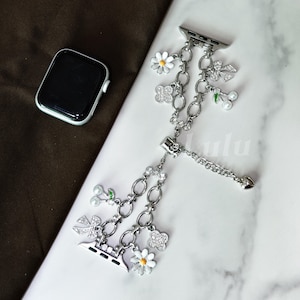 Op de afbeelding: Zilveren Apple Watch band met madelief-, kersen- en klaverbedels. De band heeft een kettingontwerp met meerdere bungelende bedels. Een zilveren Apple Watch is zichtbaar op de achtergrond.