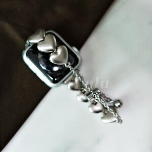 May include: Silver-toned Apple Watch band with heart-shaped charms. The band features a series of heart-shaped accents and a dangling chain of hearts. The watch face is black, and the band appears to be made of metal.