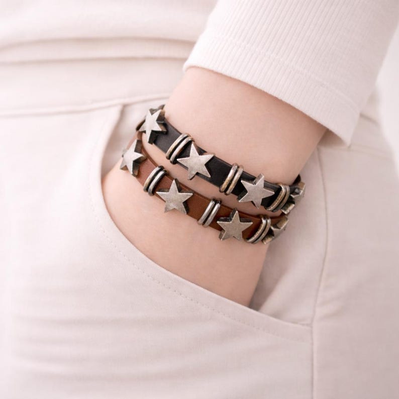 May include: Two layered bracelets featuring a black leather band and a brown leather band. Each band is adorned with metallic silver-toned star-shaped charms and small cylindrical metal spacers positioned between the stars along the length.