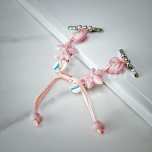May include: A pink and white charm bracelet with star and heart charms. The bracelet is made of pink string and has a silver clasp.