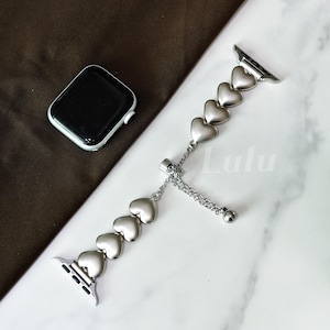 Puede incluir: Correa de Apple Watch plateada con eslabones en forma de corazón. La correa tiene una cadena y una pequeña bola para ajustar. Un Apple Watch negro es visible en el fondo.