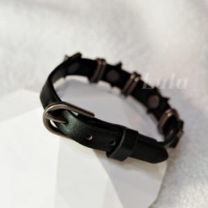 May include: A black leather-like bracelet featuring a dark metal buckle closure and a series of evenly spaced dark metal studs along the band. The word Lulu is visible in the background. The bracelet has a smooth texture and a pointed end.