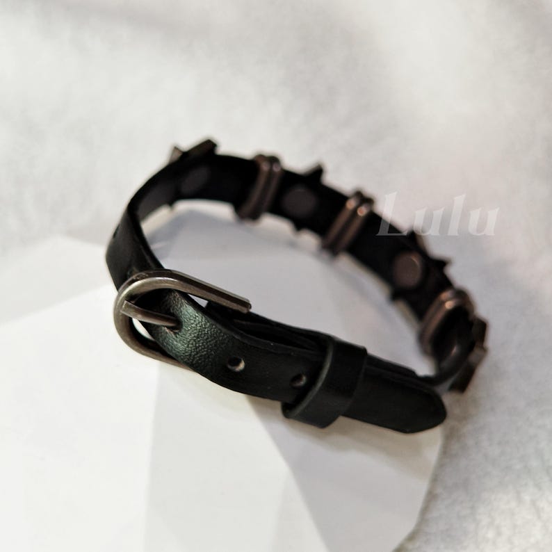 May include: A black leather-like bracelet featuring a dark metal buckle closure and a series of evenly spaced dark metal studs along the band. The word Lulu is visible in the background. The bracelet has a smooth texture and a pointed end.