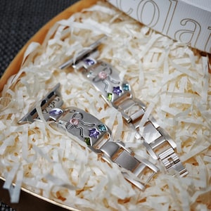 May include: A silver-toned metal watch strap featuring a matte finish and decorative cutouts. The strap is adorned with small, colourful embedded crystals in star and bow shapes, including pink, purple, green, and blue accents throughout the links.