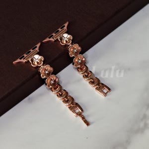 Puede incluir: Correa de reloj de oro rosa con un diseño floral y pequeños detalles brillantes. La correa está compuesta por eslabones interconectados, cada uno adornado con una flor. Tiene un cierre para una sujeción segura y está diseñada para un Apple Watch.