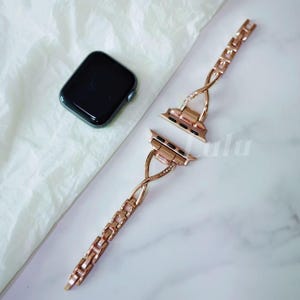 May include: Rose gold watch band with a chain link design and a black smartwatch. The band features a decorative twisted section with small crystals. The watch band is unattached, showing the connection points.