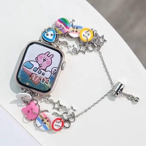 May include: A silver watch band with a colourful charm bracelet attached. The watch face shows a cartoon bunny on a plane with the time 17:03. The charms include stars, buttons, and a pink bunny.