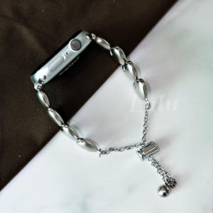 May include: Silver-toned Apple Watch band with a unique design. The band features elongated, oval-shaped links and a delicate chain with a small ball at the end. The watch face is visible, showcasing its sleek, modern design.