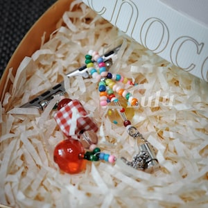 May include: A colorful beaded charm bracelet with a red and white gingham charm, a red apple charm, and a silver charm with a chain. The bracelet is in a white and brown gift box with shredded paper.