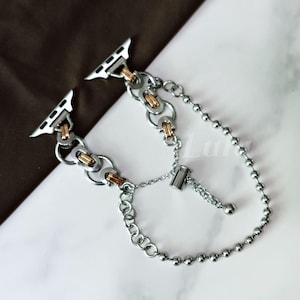 May include: Silver and gold-toned metal watch band with a chain design. The band features a series of interconnected links and a beaded chain detail. The watch band is designed to attach to a smartwatch.