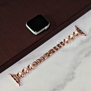 May include: A rose gold watch strap featuring flower-shaped links with small clear crystals. The strap is next to a silver smartwatch with a black screen. The watch strap is designed to fit a smartwatch.