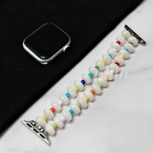 May include: White beaded Apple Watch strap with colourful accents. The strap features two strands of white beads interspersed with small, colourful beads. A silver Apple Watch is visible.