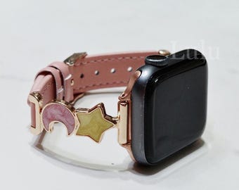 May include: A smartwatch with a light pink leather band featuring rose gold-toned metal hardware. The band is decorated with a pink crescent moon charm and a pale yellow star charm, both with a glossy finish, attached near the watch face.