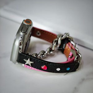 May include: A silver Apple Watch with a black and pink leather band featuring a silver chain link. The band has a star and heart design.