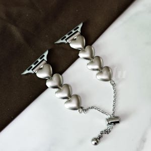 May include: Silver-toned Apple Watch band featuring a series of heart-shaped links. The band includes a chain and a small ball for adjusting the size. The band is designed to attach to the Apple Watch.