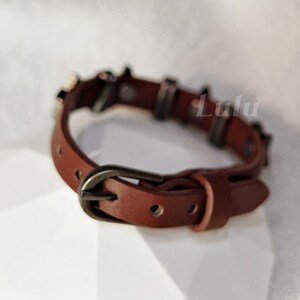 May include: A brown leather bracelet featuring a dark metal buckle and a series of dark metal studs along the band. The word Lulu is printed in white text on the right side of the bracelet. The band has multiple adjustment holes for sizing.