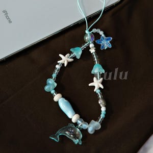 May include: A beaded phone charm featuring a dolphin, starfish, and heart-shaped beads in shades of blue and white. The charm includes the text "lulu" and is attached to a light blue cord.