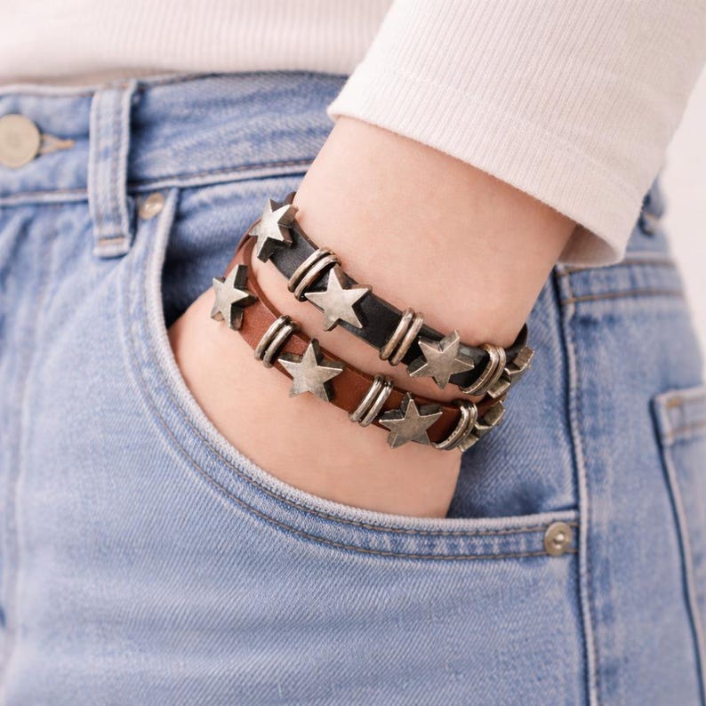 May include: Two leather bracelets, one black and one brown, each featuring a series of metallic silver-toned star-shaped charms and small cylindrical metal spacers arranged along the length of the bands.
