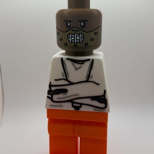 May include: A custom Lego figure depicting a character with a brown head and mask, white torso with arms crossed, and orange pants. The figure is a unique, handcrafted item.