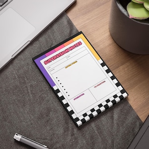May include: A rectangular notepad with the words "SUPERVISION NOTES" in pink and orange. The notepad has a black and white checkered border and sections for notes, action items, and to-do lists. The notepad is on a desk.