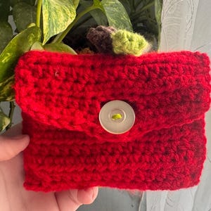 May include: A handmade, red crochet wallet with a flap closure and a large, round, off-white button. The wallet is made of a thick, textured yarn and appears to be sturdy. The background includes green foliage and a sheer curtain.