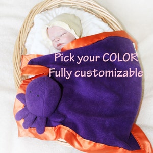 May include: A purple fleece baby blanket with an orange satin trim and a purple octopus plush toy. The blanket is in a wicker basket with a baby wearing a white hat.