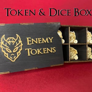 May include: Black wooden token and dice box with gold trim and the text "Token & Dice Box". The box is open, revealing compartments with enemy tokens. The lid features a gold graphic and the words "Enemy Tokens".