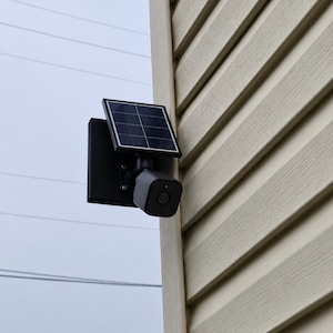 May include: A black security camera mounted on a wall, featuring a square solar panel attached to the top. The camera has a rounded square housing with a central lens and is positioned on the corner of a building with light beige horizontal siding.