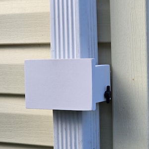 May include: A white rectangular object attached to a white downspout with a black knob. The downspout is mounted on a light yellow siding. The object appears to be a gutter accessory.