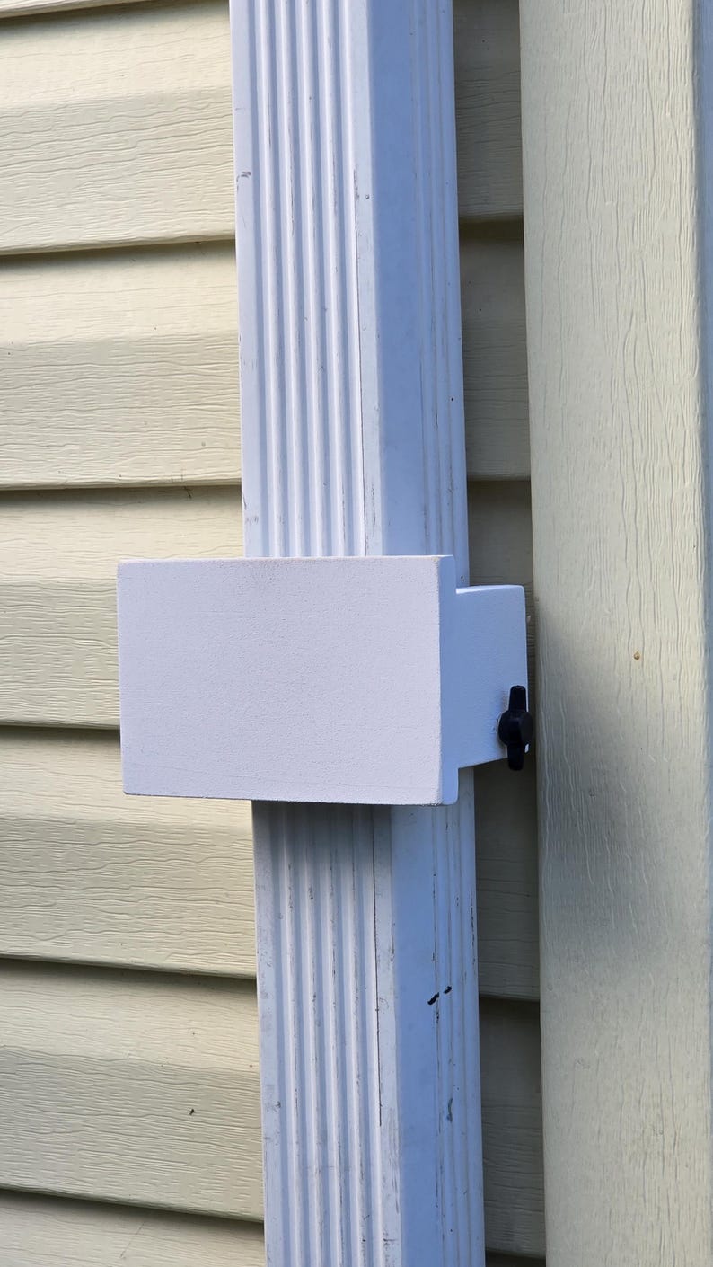 May include: A white rectangular object attached to a white downspout with a black knob. The downspout is mounted on a light yellow siding. The object appears to be a gutter accessory.