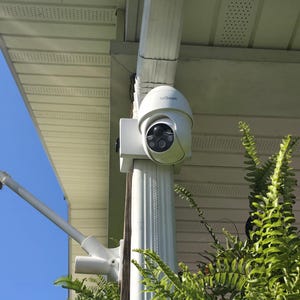 May include: A white outdoor security camera mounted on a white column, positioned under a white eave and gutter. The camera has a dome shape with the brand name "ieGeek" visible. Green ferns and a blue sky are in the background.