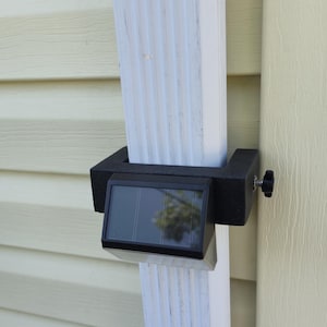 May include: A black solar-powered gutter light is mounted on a white gutter. The light has a rectangular shape with a clear panel and is attached with a black bracket and a black knob.