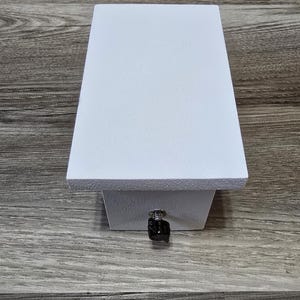 May include: A white, rectangular box with a textured surface. The box has a small, black knob on the front. The box is sitting on a wooden surface.