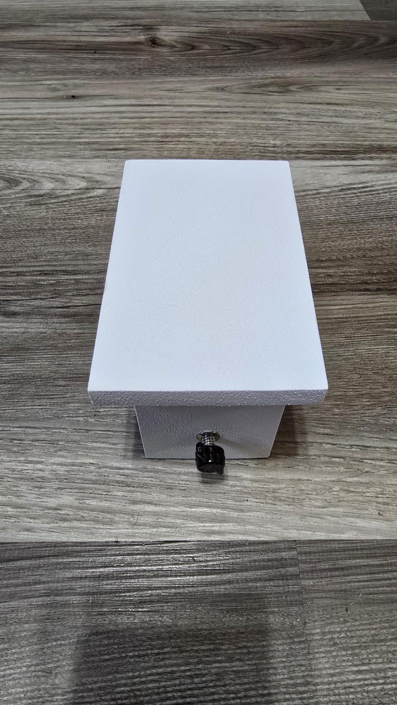 May include: A white, rectangular box with a textured surface. The box has a small, black knob on the front. The box is sitting on a wooden surface.