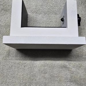 May include: A white, U-shaped object with a flat base and two vertical supports. A black knob is visible on the right side. The object is sitting on a gray, textured surface.
