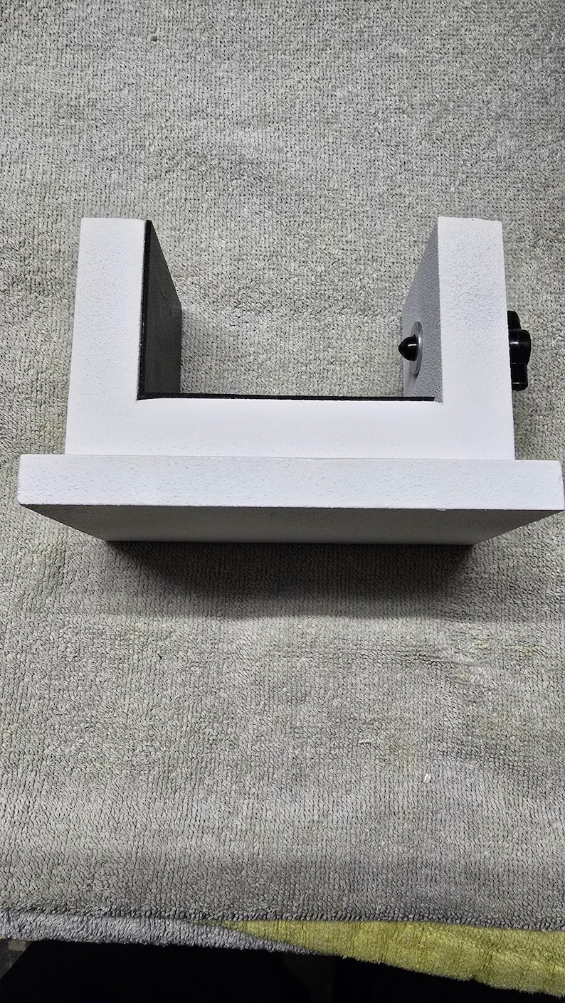 May include: A white, U-shaped object with a flat base and two vertical supports. A black knob is visible on the right side. The object is sitting on a gray, textured surface.