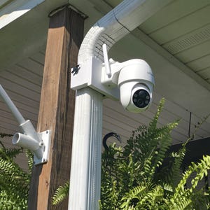 May include: White outdoor security camera mounted on a white pole, with a black lens and antenna. The camera is positioned near a wooden post and green foliage, with a white gutter above.