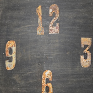 May include: A rustic clock face with a dark gray wooden background and a light brown wooden frame. The numbers 12, 3, 6, and 9 are displayed in a distressed, rusty metal font.