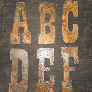 Puede incluir: Seis letras metálicas, A, B, C, D, E y F, con un aspecto oxidado y envejecido. Las letras están cortadas de metal y tienen un aspecto oscuro y desgastado, con tonos marrones y anaranjados de óxido.