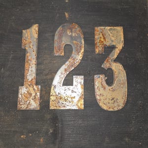 May include: A wooden sign displaying the numbers 1, 2, and 3. The numbers are crafted from aged metal with a rusty appearance, set against a dark stained wooden backdrop. The sign is framed with natural wood.