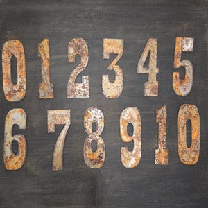Puede incluir: Números de metal oxidado, del cero al diez, dispuestos sobre un fondo de madera gris oscuro. Los números presentan un aspecto envejecido con tonos naranja y marrón, enmarcados en un marco de madera.