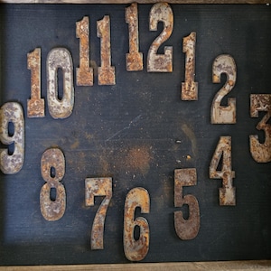 May include: A rustic clock face with metal numbers arranged on a dark background within a wooden frame. The numbers, from 1 to 12, have a weathered, rusty appearance, suggesting an antique or vintage aesthetic. The clock face is a decorative item.