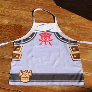 Op de afbeelding: Een lichtblauwe schort met een zak en een witte nekband. De schort heeft een rood-wit grafisch ontwerp in het midden, met bruine en gouden accenten. De schort is ontworpen voor gebruik in de keuken.